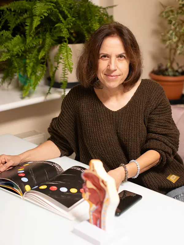 Kobieta siedzi przy stole z książką do anatomii i modelem, otoczona roślinami.