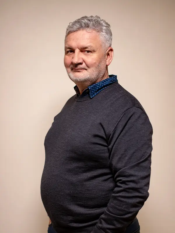 Mężczyzna o siwych włosach i brodzie, ubrany w ciemny sweter, stoi na jednolitym tle, zwrócony twarzą do kamery.