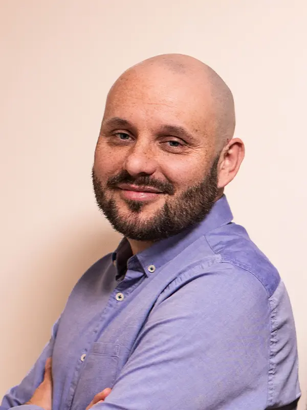 Łysy mężczyzna z brodą, w niebieskiej koszuli, uśmiechnięty, pozuje na jednolitym tle.