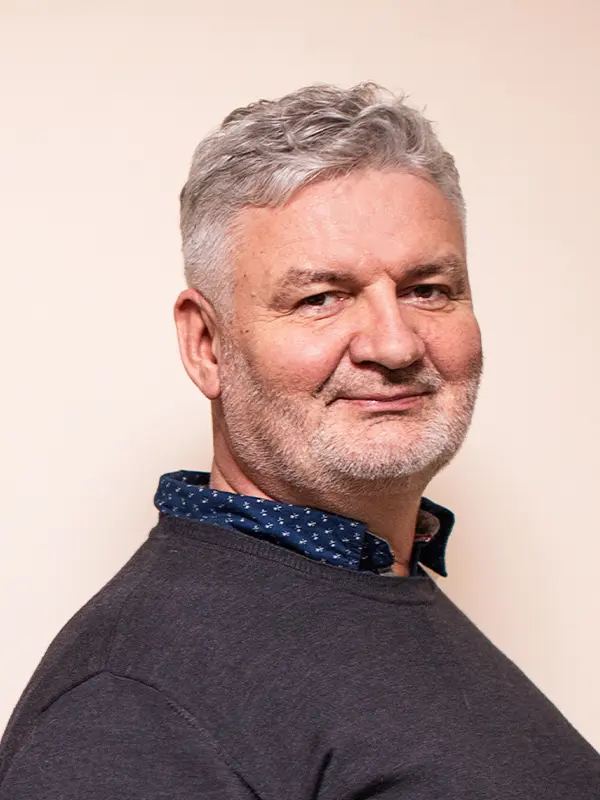 Starszy mężczyzna z siwymi włosami i brodą, ubrany w ciemny sweter, delikatnie się uśmiechający na jednolitym tle.