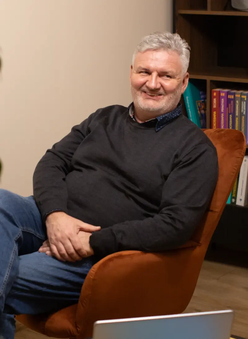 Uśmiechnięty mężczyzna o siwych włosach siedzi na pomarańczowym krześle obok regału z książkami.