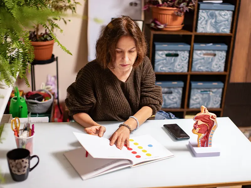 Kobieta czyta kolorową książkę przy stole, obok którego znajduje się model ludzkiego gardła.