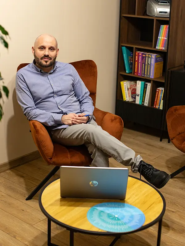 Łysy mężczyzna w niebieskiej koszuli siedzi na krześle obok małego stolika z laptopem, w tle widać półkę z książkami.