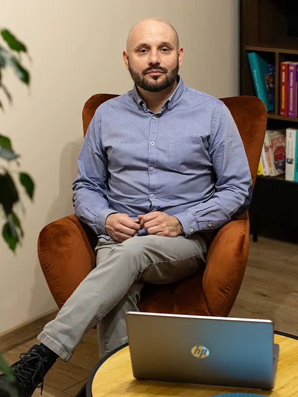 Osoba z brodą siedzi na pomarańczowym krześle obok laptopa, za nią na półce widać książki.