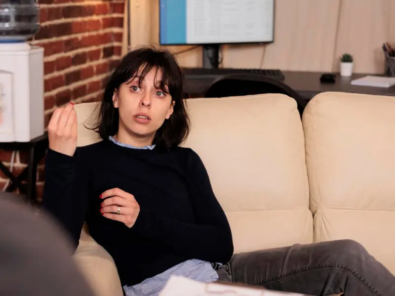 Kobieta siedzi na kanapie, gestykulując ręką, w pokoju z odsłoniętą ścianą z cegły i biurkiem komputerowym w tle.