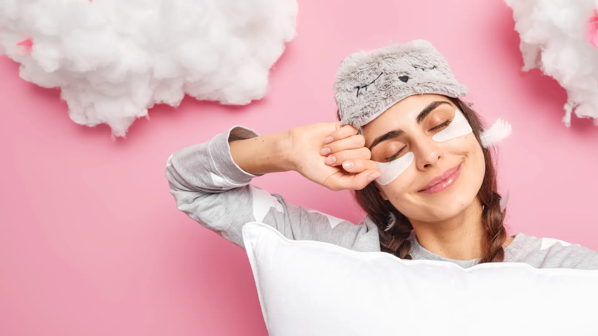 Uśmiechnięta kobieta z zakrytymi oczami i maską do spania, trzymająca poduszkę, na różowym tle z puszystymi chmurkami.