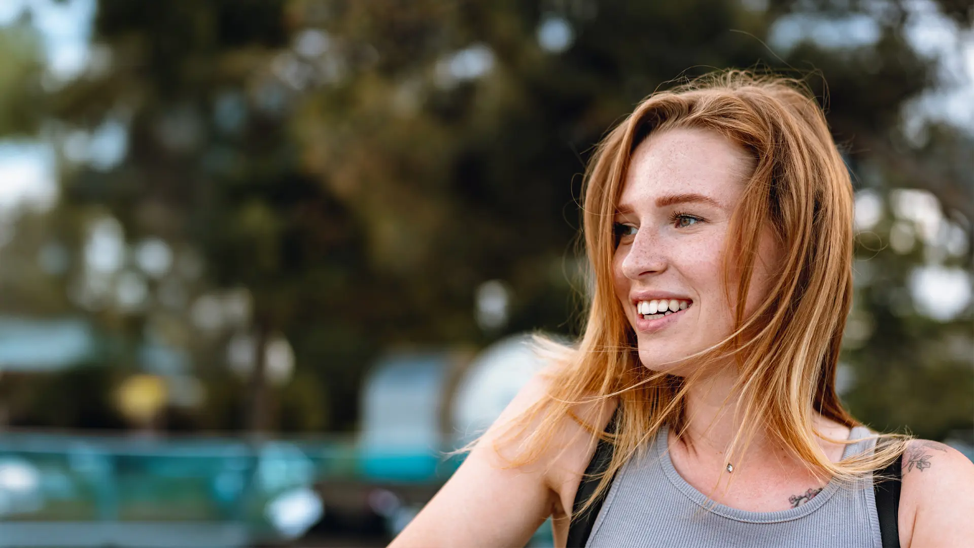 Uśmiechnięta kobieta o rudych włosach stoi na zewnątrz, ubrana w szary podkoszulek, na tle rozmazanych drzew.