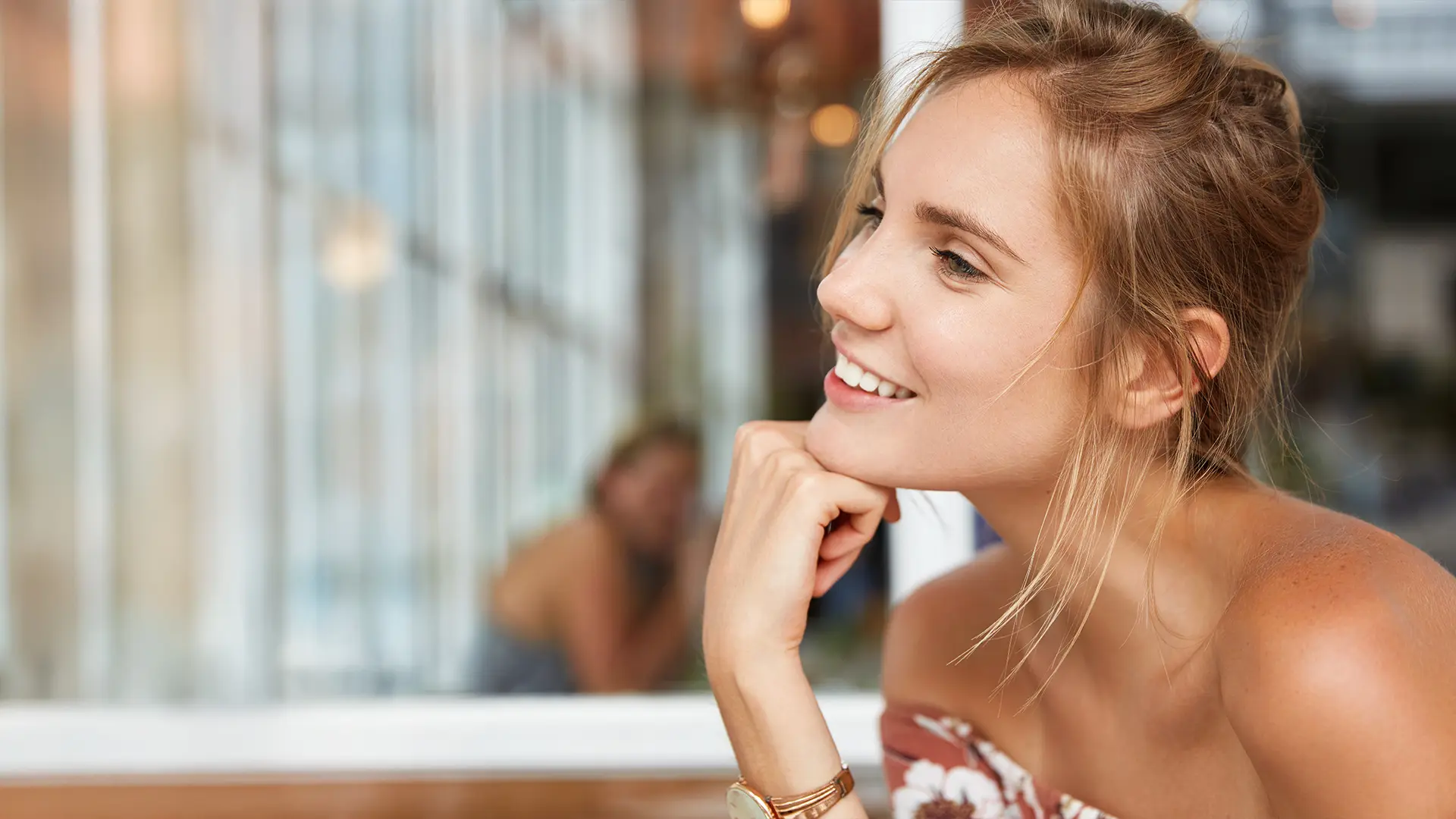 Uśmiechnięta kobieta siedzi przy oknie, z brodą opartą na dłoni, ubrana w kwiecistą bluzkę z odkrytymi ramionami.