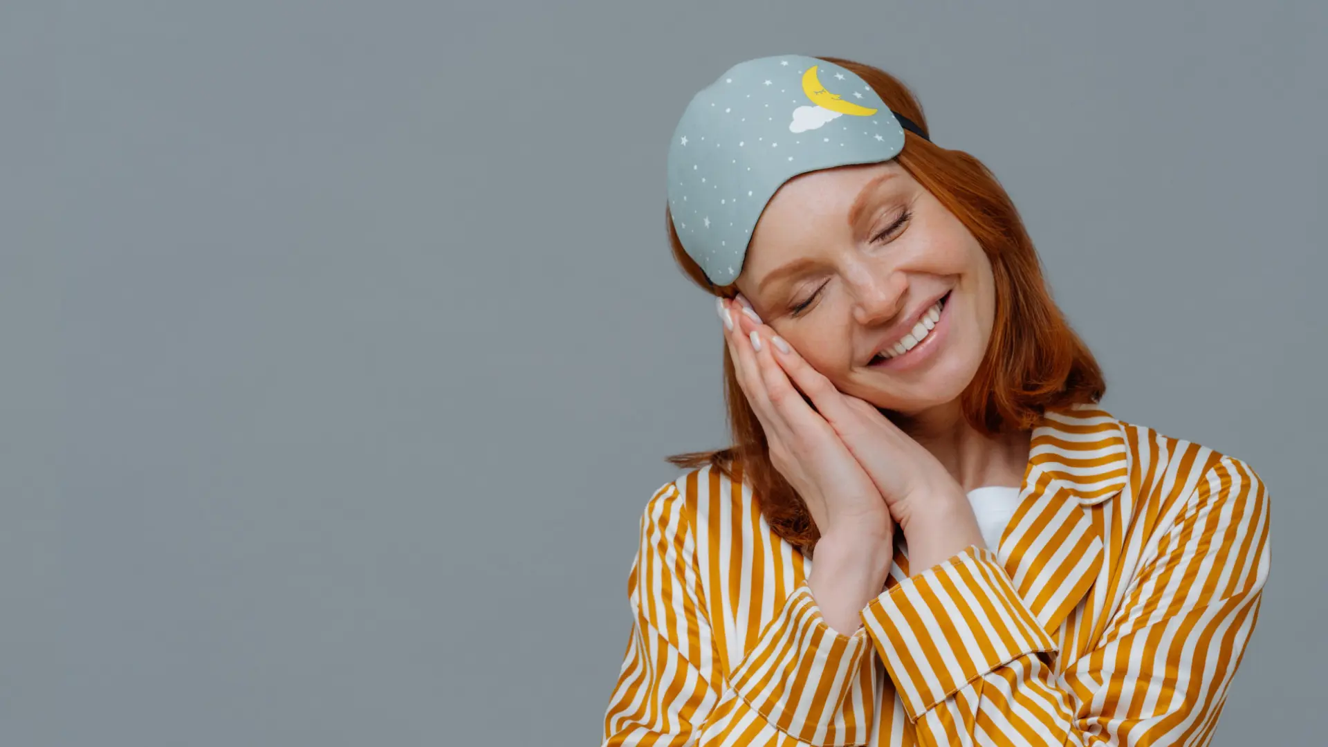 Kobieta w pasiastej piżamie i masce do spania uśmiecha się, trzymając dłonie pod policzkami, jakby spała, na szarym tle.
